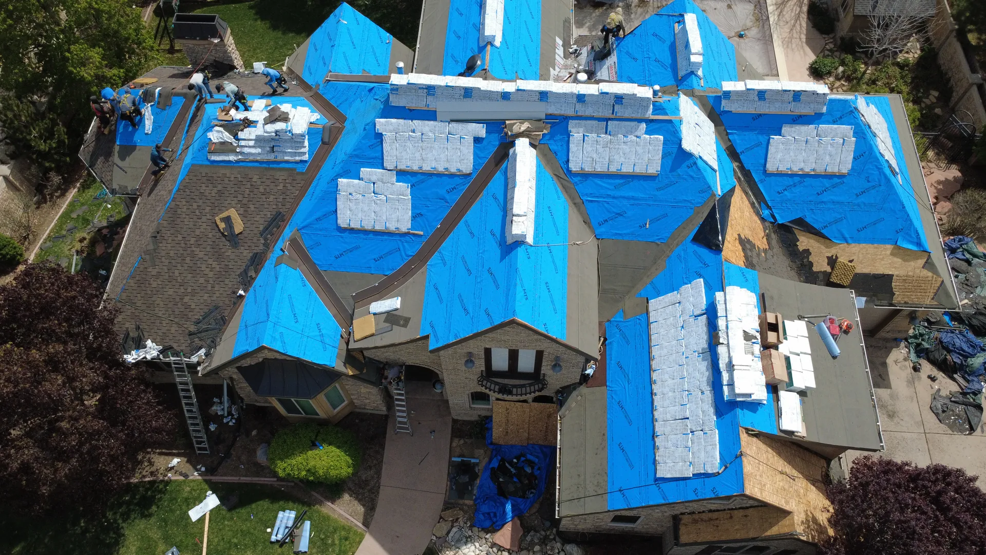 Top-down drone photograph of the same Heber Valley custom home roof showing packaged shingle bundles distributed across every plane of the roof on top of blue synthetic underlayment, with Frame Restoration Utah crew members visible at work on the ridgeline.