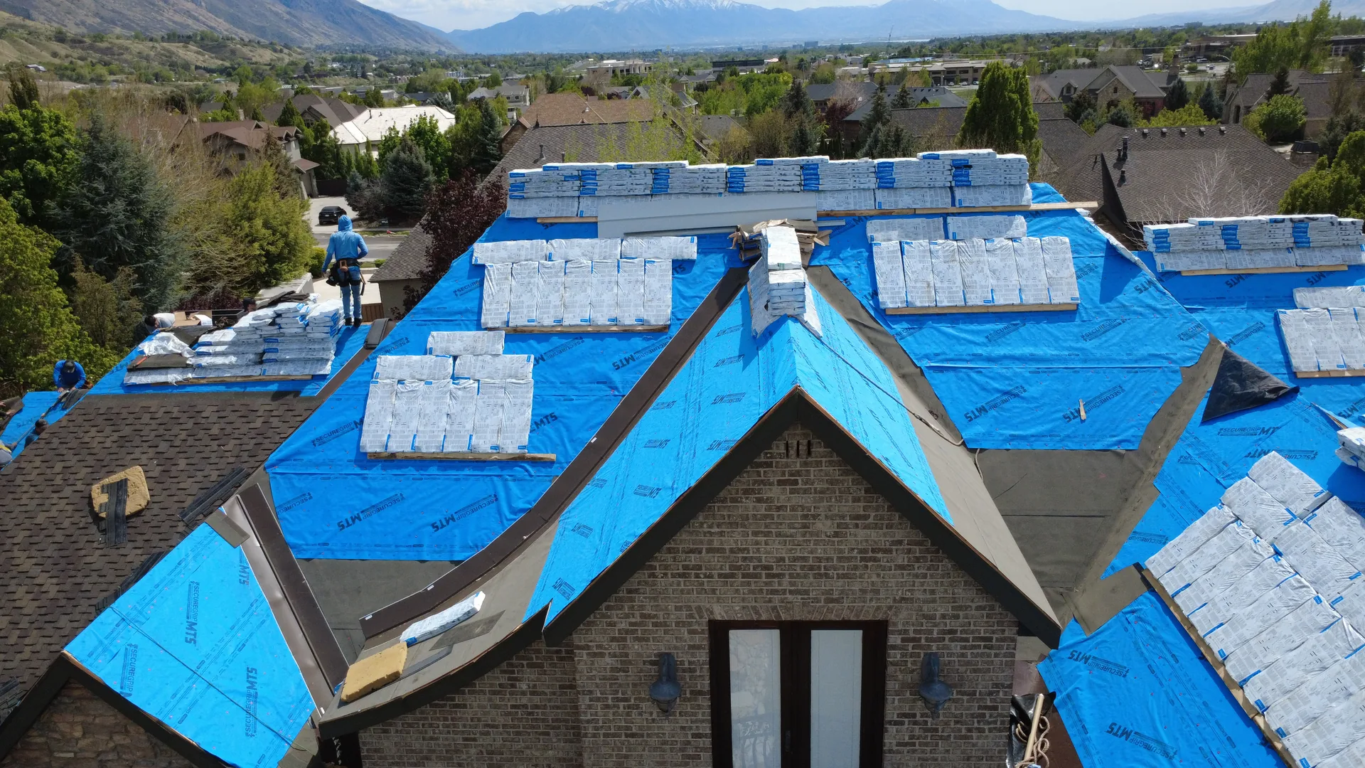 Angled aerial drone photograph of the Heber Valley custom home reroof with a Frame Restoration Utah crew member installing architectural shingles on a dormer, the Wasatch Front mountain range visible in the distance.