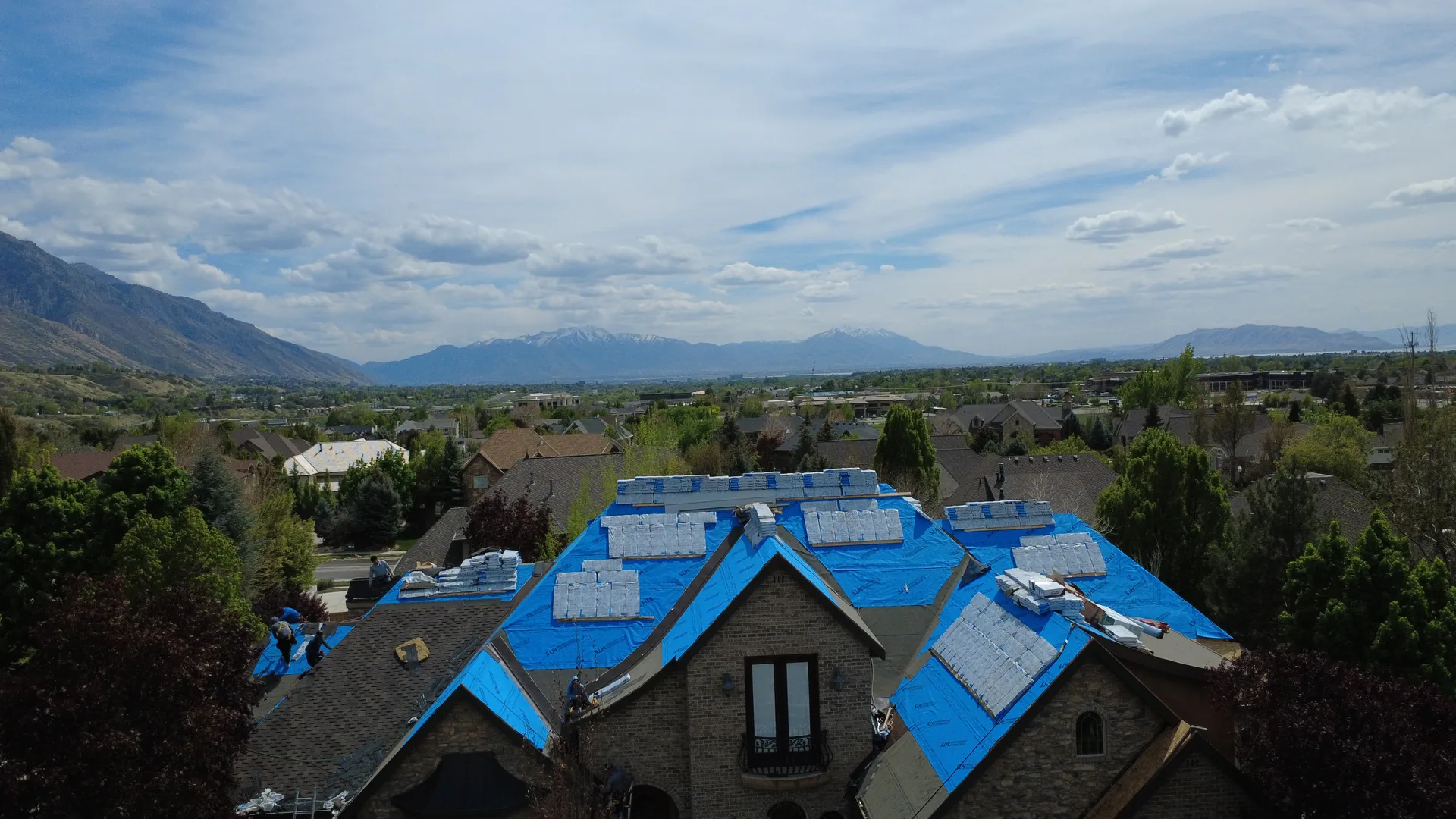 Aerial drone photograph of a Heber Valley custom home mid-reroof by Frame Restoration Utah — blue Suntuff synthetic underlayment fully laid, packaged architectural shingle bundles staged in stacks across every roof plane, Wasatch Back mountain ridge and valley visible in the background under a blue sky.