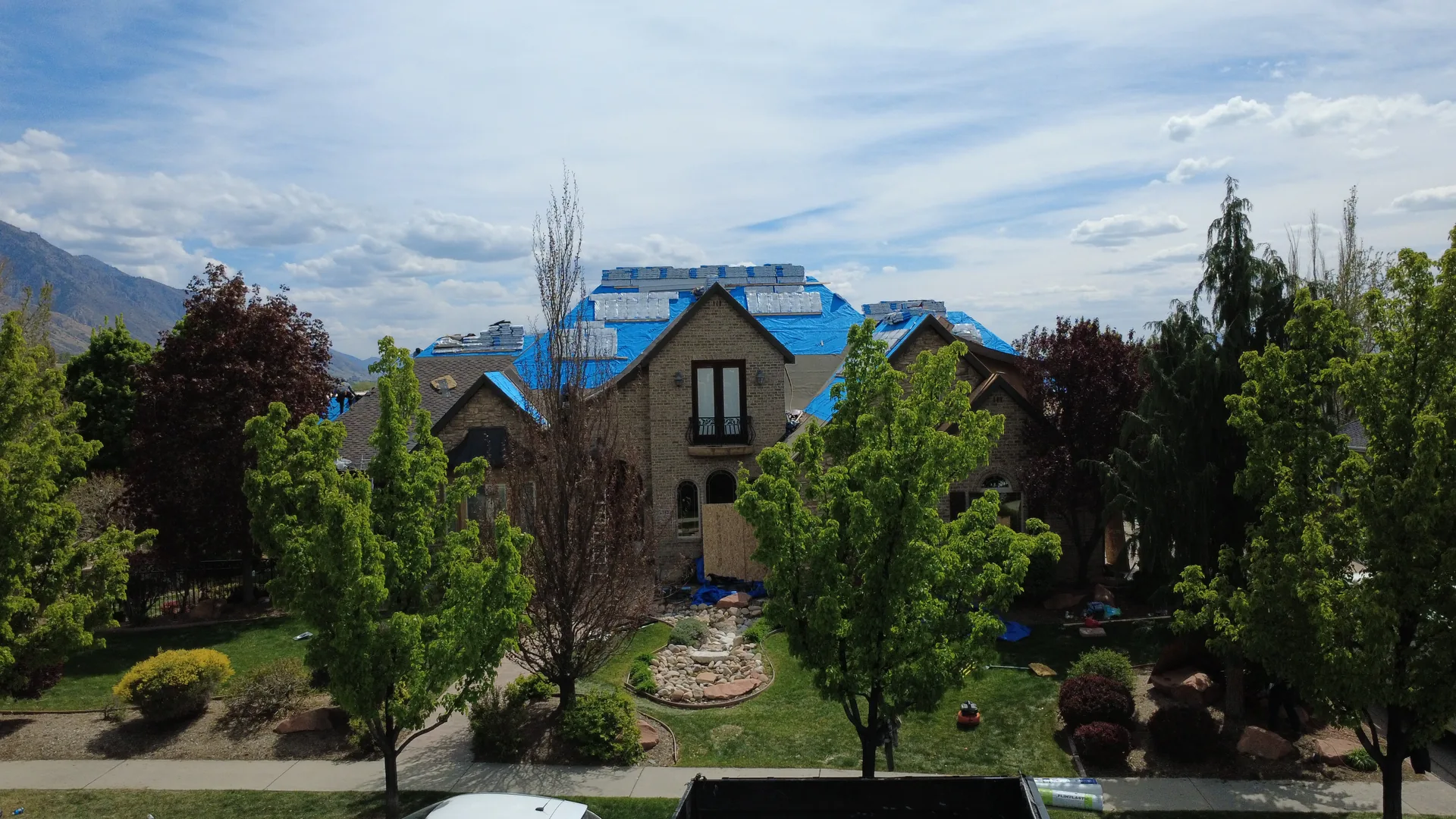 Drone photograph of the same Heber Valley custom home reroof taken from the front yard elevation, showing the two-story brick-and-stone elevation with the blue synthetic underlayment visible on the roof and the Frame Restoration Utah roll-off dumpster on the driveway.